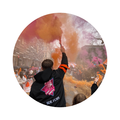 Photo d'un étudiant de dos brandissant un fumigène orange lors d'un rassemblement festif en extérieur, entouré d'une foule et de fumées colorées.
