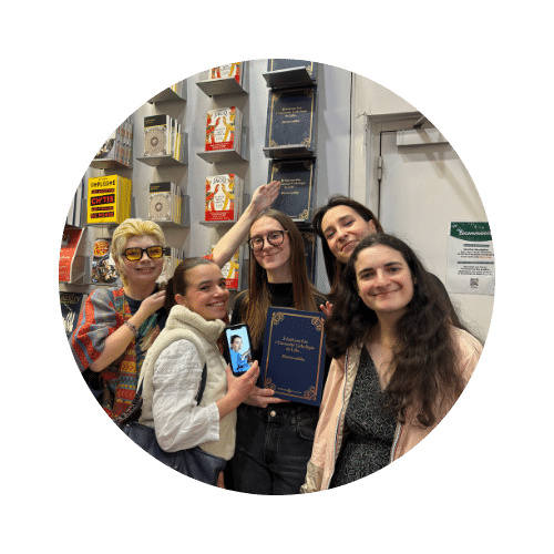 Groupe de cinq jeunes femmes souriantes dans une librairie, l'une d'elles tenant un livre à couverture bleue et un smartphone.