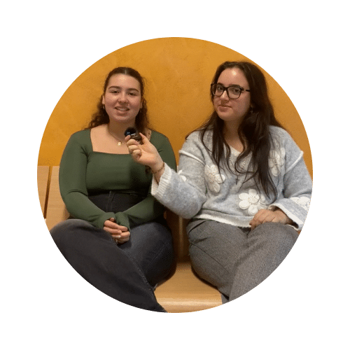Deux jeunes femmes assises côte à côte sur un banc en bois. Celle de droite, portant des lunettes et un pull gris avec des motifs floraux, tient un microphone vers la jeune femme de gauche, qui porte un haut vert et sourit.