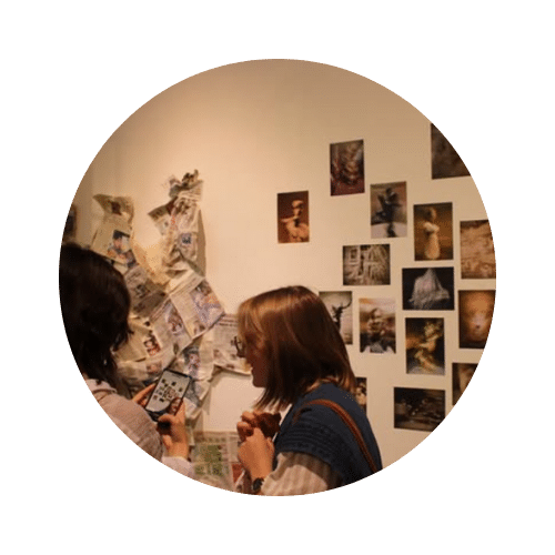 Deux personnes de profil observant une exposition murale. À gauche, un assemblage de papiers et de journaux déchirés est fixé au mur ; à droite, une série de photographies artistiques est disposée de manière régulière. L'une des personnes tient un smartphone pour prendre une photo.