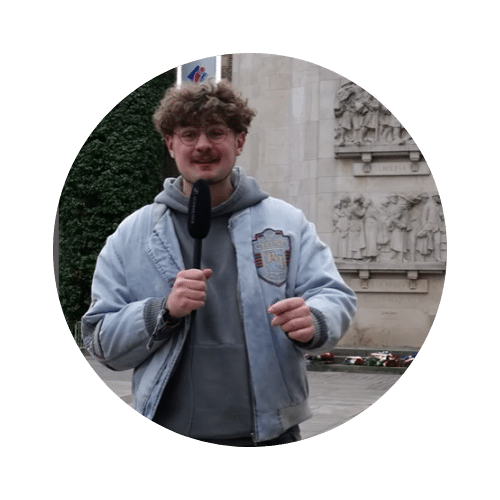 Un jeune homme avec des lunettes et des cheveux bouclés, portant une veste en jean bleu sur un sweat à capuche gris, tenant un microphone. Il se tient devant un mur en pierre sculpté de bas-reliefs classiques.