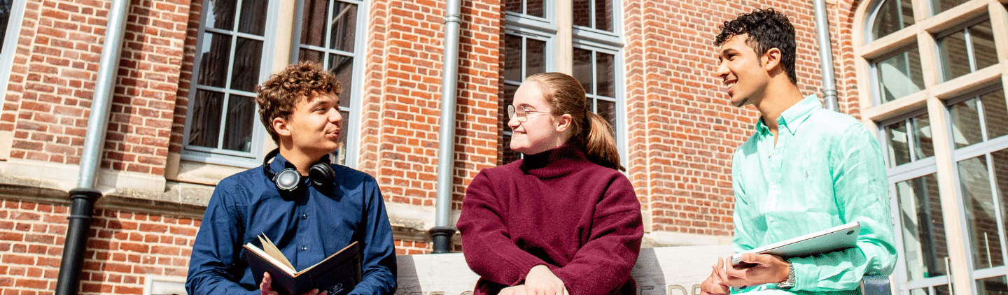 Trois étudiants, deux jeunes hommes et une jeune femme, discutent et sourient ensemble sur un banc devant un bâtiment universitaire en briques rouges.