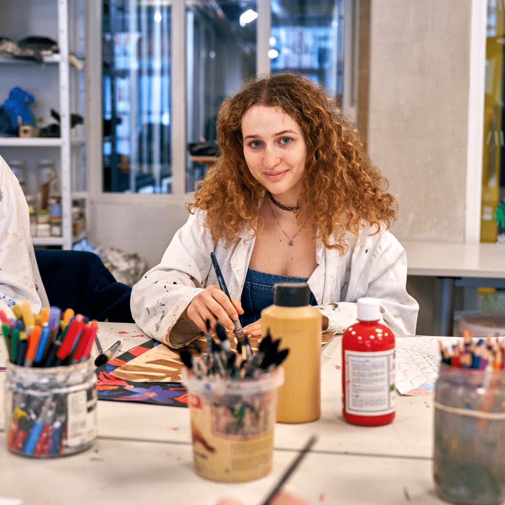 Une étudiante en blouse blanche travaillant sur une œuvre artistique dans un atelier, entourée de pinceaux et de pots de peinture.
