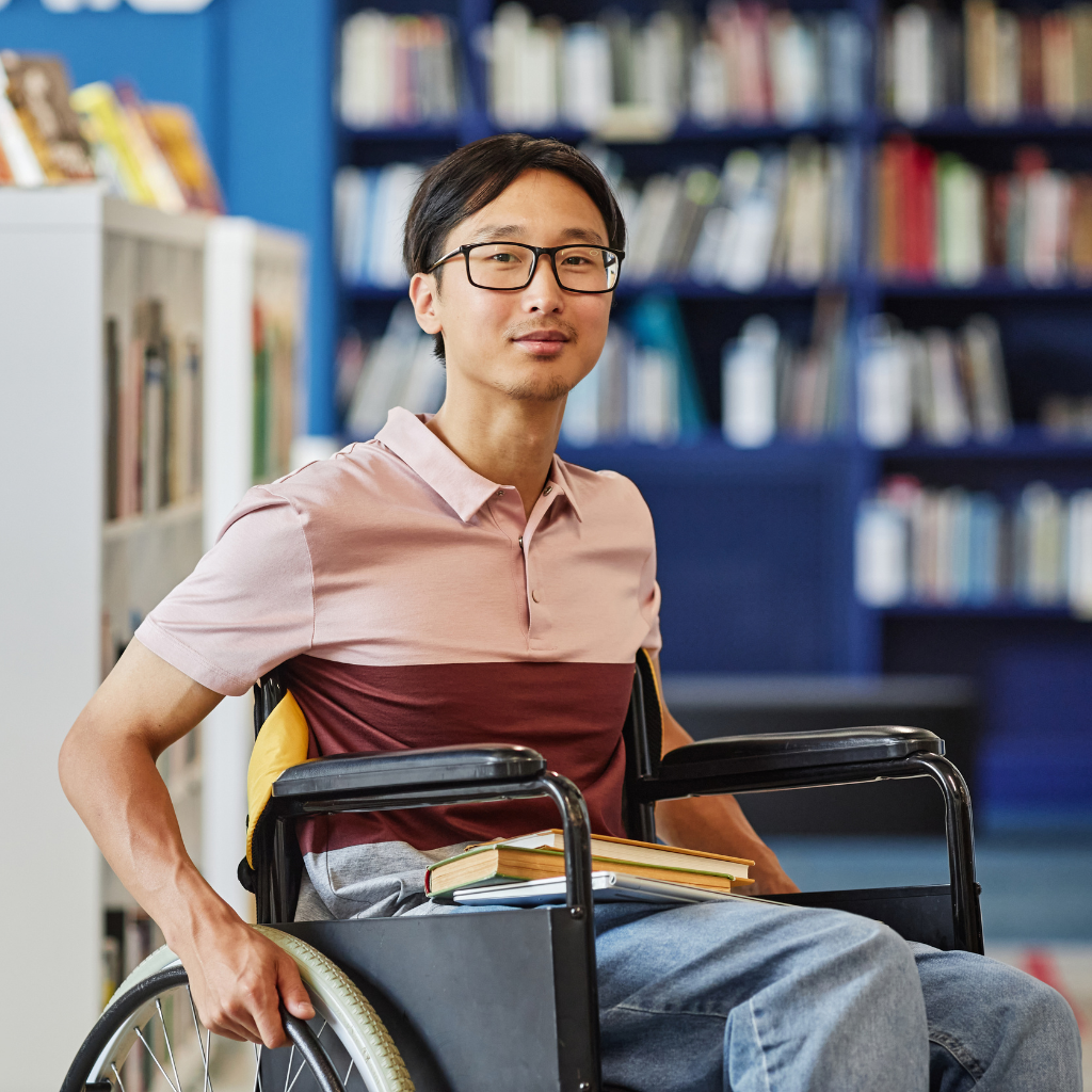 Un étudiant en fauteuil roulant souriant dans une bibliothèque, illustrant les valeurs d'inclusion et l'engagement pour l'égalité des chances au sein de l'établissement.
