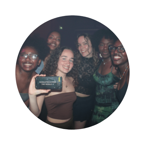 Groupe de jeunes femmes souriantes lors d'une soirée, l'une d'elles tenant un téléphone affichant le texte « SUMAAFRICA OVER THE WORLD