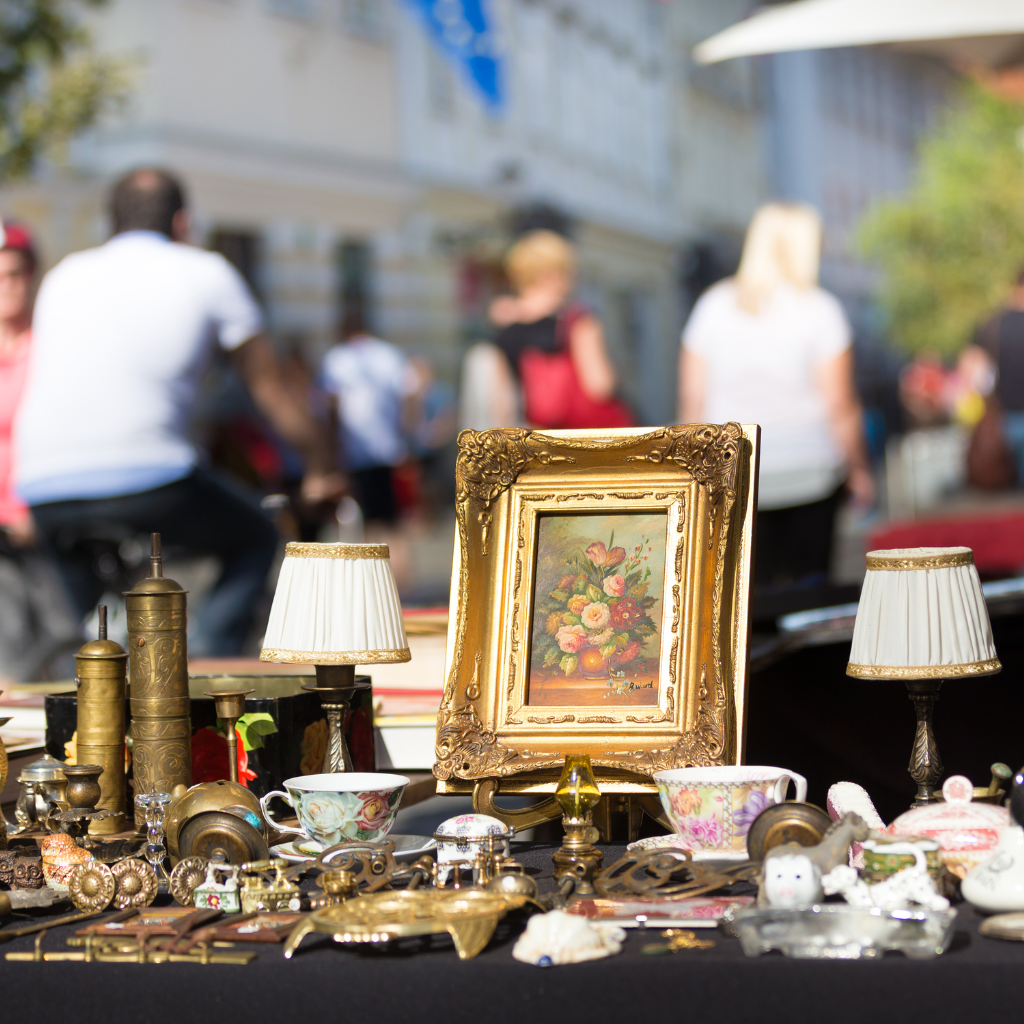Outdoor flea market table featuring a small gold-framed floral painting, porcelain cups, and vintage lamps, with blurred pedestrians in the background.