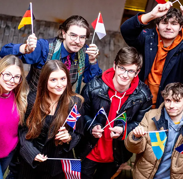 Un groupe de six jeunes étudiants souriants posant ensemble, chacun tenant un petit drapeau national. On reconnaît notamment les drapeaux de l'Allemagne, de l'Autriche, du Royaume-Uni, des États-Unis, de l'Afrique du Sud et de la Suède. Ils portent des vêtements variés (vestes en cuir, pulls, blazers), évoquant un environnement universitaire multiculturel.